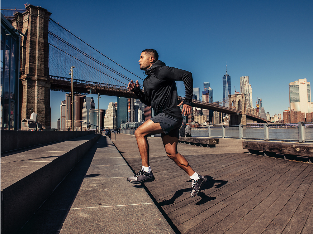A person in athletic attire runs up steps near a river, with a city skyline and bridges visible in the background under a clear blue sky.
