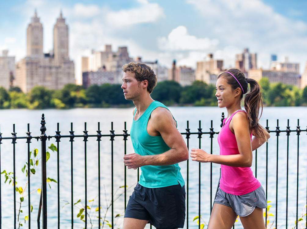 Two people jog along a waterfront path with city buildings in the background. They wear athletic attire and pass by a black metal fence on a clear day.