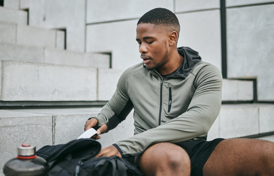 A man sits on concrete steps, wearing a green hoodie and black shorts, looking into his black backpack. A water bottle is nearby.