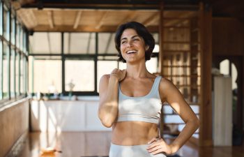 A woman in athletic wear stands smiling indoors, with natural light streaming through large windows.