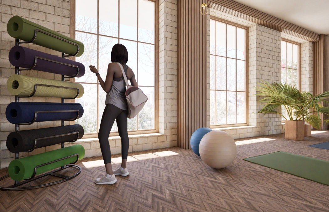 Person with a bag stands near a rack of rolled yoga mats in a well-lit room with large windows, wooden floors, and potted plants.