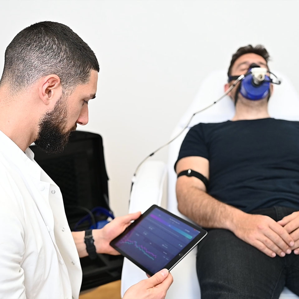 A man lies on a medical chair using a breathing mask, while a healthcare professional monitors data on a tablet.