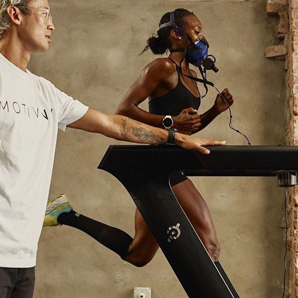 A woman runs on a treadmill wearing a breathing mask, while a man stands beside her, observing and holding the treadmill.